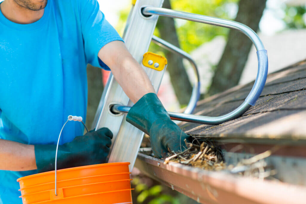 Closeup view of an anonymous worker standing on a ladder leaning on the roof of a house and scooping out the eavestrough or roof gutters to clean them for the upcoming winter. He is wearing protective gloves and scooping the collection into a bucket.